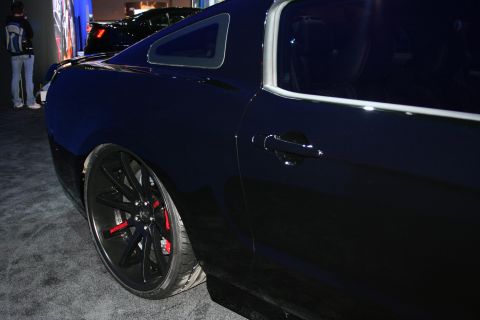 1998 Ford Crown Victoria - Low side view of a dark Ford Mustang coupe showing a black multi-spoke wheel with a red brake caliper at a dim show.