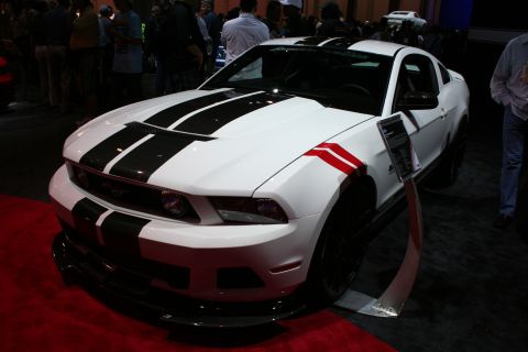 1998 Ford Crown Victoria - white Ford Mustang coupe with black center stripes and a red hash-mark fender accent on red carpet at an indoor show.