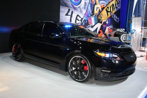 1998 Ford Crown Victoria - blacked-out Ford Taurus sedan with dark wheels and red brake calipers sits on a white stage before a graffiti-style backdrop.