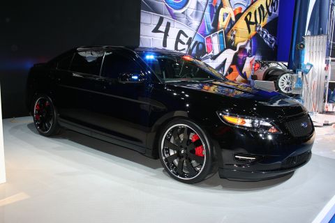 1998 Ford Crown Victoria - Front three-quarter of a blacked-out Ford Taurus sedan with dark wheels and red calipers on a white stage with a graffiti backdrop.