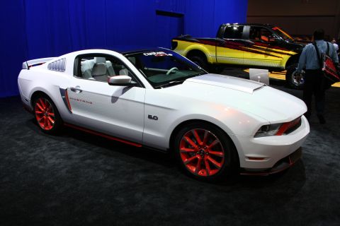 1998 Ford Crown Victoria - white Ford Mustang 5.0 coupe with red side stripes and red-painted wheels displayed on an auto show floor.