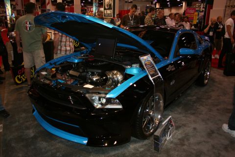 1998 Ford Crown Victoria - black Ford Mustang with bright blue accents and an open hood revealing a supercharged engine, displayed at a show.