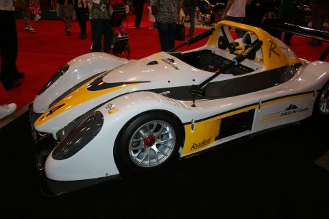 1998 Ford Crown Victoria - white and yellow open-cockpit Radical-style race car with roll hoop and exposed cockpit displayed on a show floor.