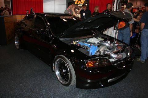 1998 Ford Crown Victoria - black modified sedan sits on display with its carbon-fiber hood open, revealing a polished turbocharged engine bay.