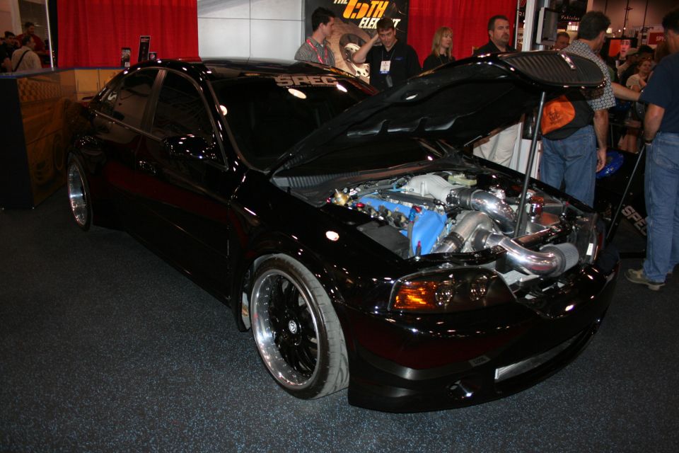 Black tuned sedan with carbon hood open showing turbocharged engine at indoor show