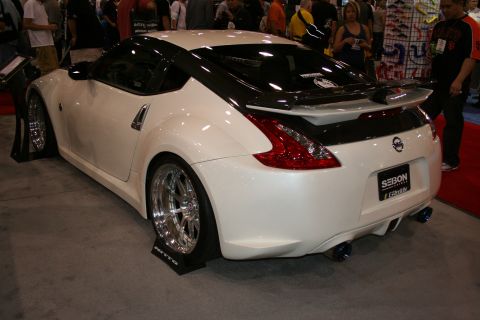 1998 Ford Crown Victoria - white Nissan 370Z with a carbon-fiber hood and roof shown from the rear three-quarter at an indoor show.