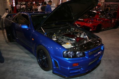 1998 Ford Crown Victoria - blue Nissan Skyline GT-R R34 sits with its hood open, displaying the engine bay at an indoor show.