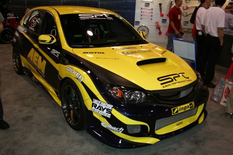 1998 Ford Crown Victoria - yellow and purple widebody Subaru WRX STI hatchback covered in sponsor decals on a show floor.