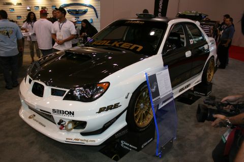 1998 Ford Crown Victoria - white Subaru Impreza WRX STI sedan with a carbon-fiber hood, gold wheels and Falken decals at an indoor show.