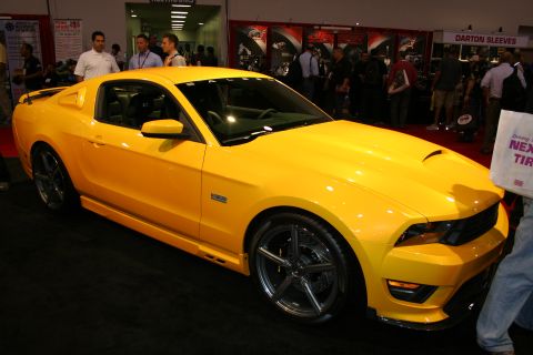 1998 Ford Crown Victoria - yellow Ford Mustang coupe with Saleen styling sits on display at a busy indoor automotive trade show.