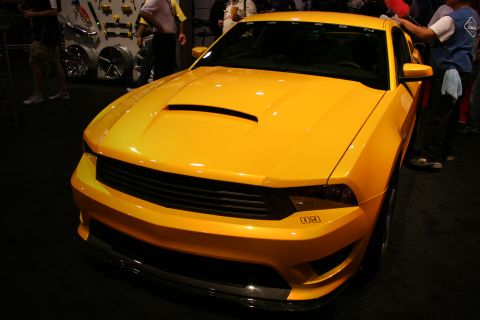 1998 Ford Crown Victoria - Front view of a yellow Saleen Mustang with a hood scoop and lowered stance, displayed near a wheel wall.