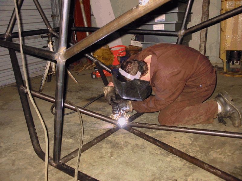 Welder in brown coveralls welding a joint on a bare steel tubular roll cage in a shop