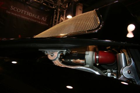 1998 Ford Crown Victoria - gold wire-mesh hood scoop sits atop a black vehicle with a 'Scottsdale, AZ' collector auction banner in the background.