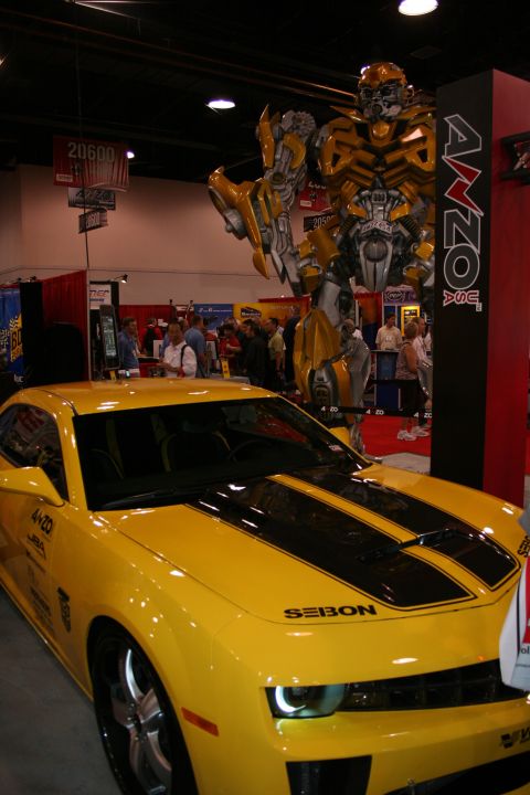 1998 Ford Crown Victoria - yellow coupe with black racing stripes parked beside a tall yellow robot statue at an indoor trade-show booth.