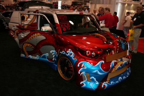 1998 Ford Crown Victoria - boxy compact car wrapped in custom red, silver, octopus-tentacle and ocean-wave artwork on display at an indoor show.