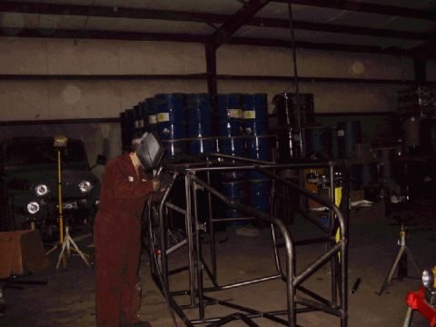 1994 Ford Bronco - A person welds a metal frame in a workshop, with a dark SUV and stacked barrels visible.