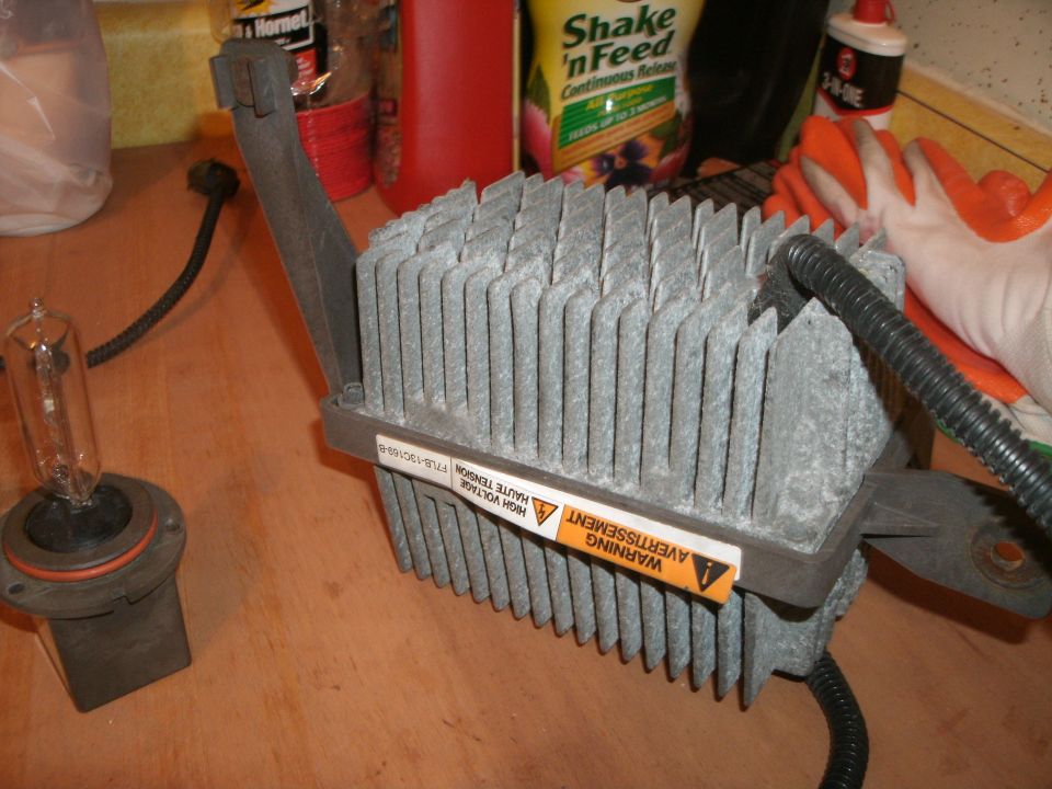 Headlight ballast with fins and a separate bulb on a wooden surface. Warning label visible