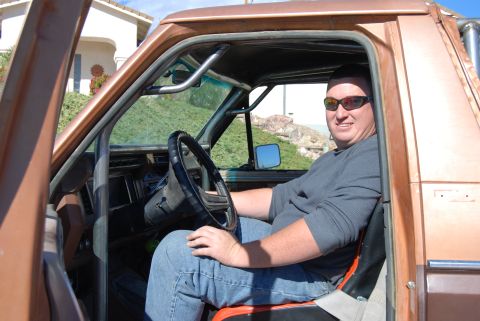 1982 Ford Bronco - Man sits in the driver's seat of a modified brown SUV, showing the interior and roll cage.