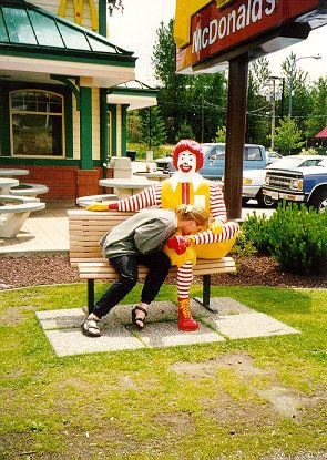 1977 Ford Mustang II - A person interacts with a Ronald McDonald statue outside a McDonald's restaurant, with vehicles visible in the background.