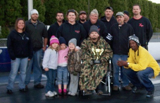 Group of people, including children and a man in a wheelchair holding a trophy, pose for a photo at a drag strip