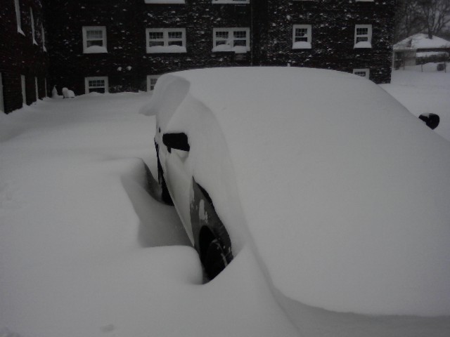 Car almost completely covered in deep snow, only a small portion of the side and a wheel are visible