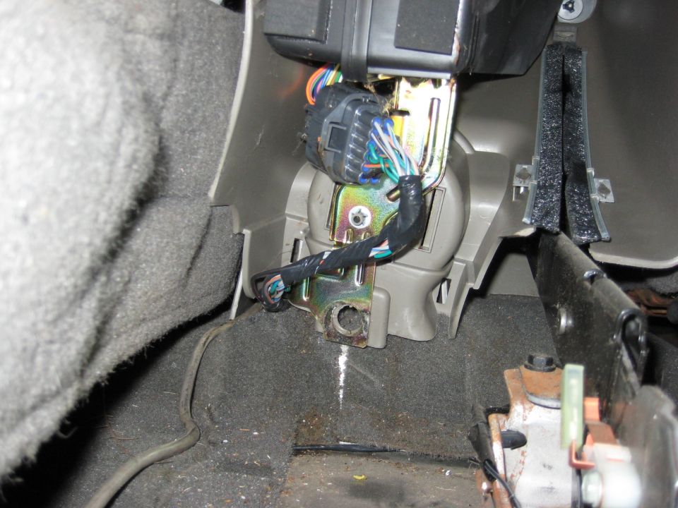 Wiring harness connected to a metal bracket inside a vehicle's interior, with gray carpet and plastic trim visible