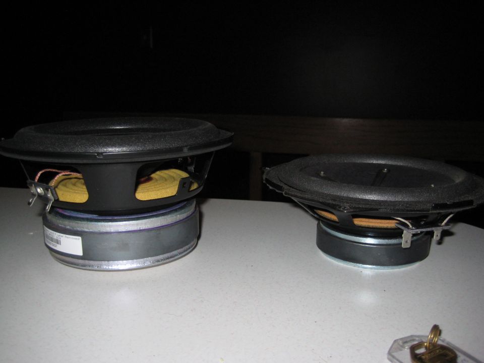Two car speakers on a white surface, one larger than the other. A label is visible on the base of the larger speaker