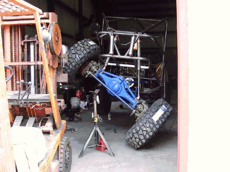Tube-frame buggy with blue front axle at full articulation on a jack stand in a shop
