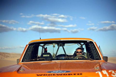 1986 Ford Bronco - Driver in an orange off-road vehicle with a roll cage, seen from the front with a desert landscape in the background.