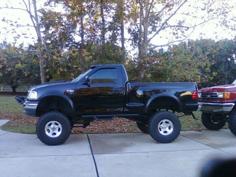 2000 Ford F-150 - Black tenth-gen Ford F-150 single-cab pickup with suspension lift and oversized tires parked on grass next to a red truck.