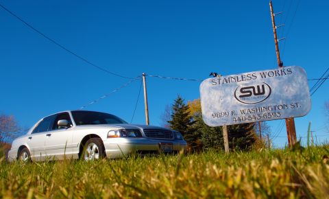 1998 Ford Crown Victoria - silver four-door sedan parked in grass beside a 'Stainless Works' shop sign under a clear blue sky.