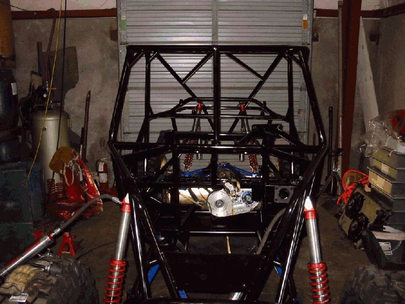 Bare black tube-chassis buggy frame in garage with red-spring coilovers and exposed transfer case