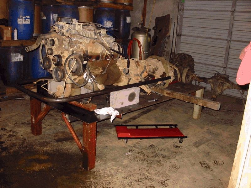 Pulled V engine and transmission on a shop bench with bent black tube chassis rail and red creeper