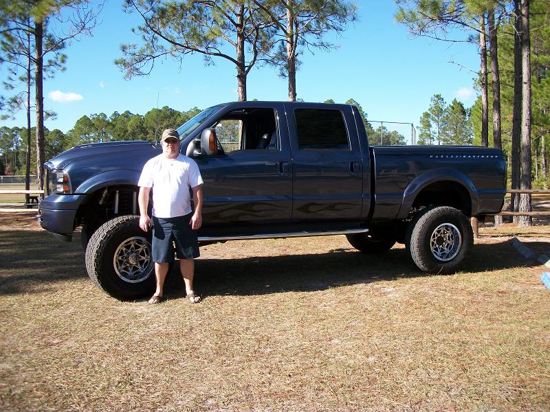 Man in white shirt and shorts stands beside a dark blue lifted Ford F-250 Super Duty pickup truck with flame decals