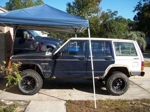 1992 Jeep Cherokee - A two-tone Jeep Cherokee is parked under a canopy, with a pickup truck partially visible behind it.