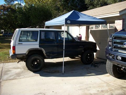 1992 Jeep Cherokee - A side view of a two-tone Jeep Cherokee parked under a blue canopy, with a portion of a blue Ford truck visible to its right.