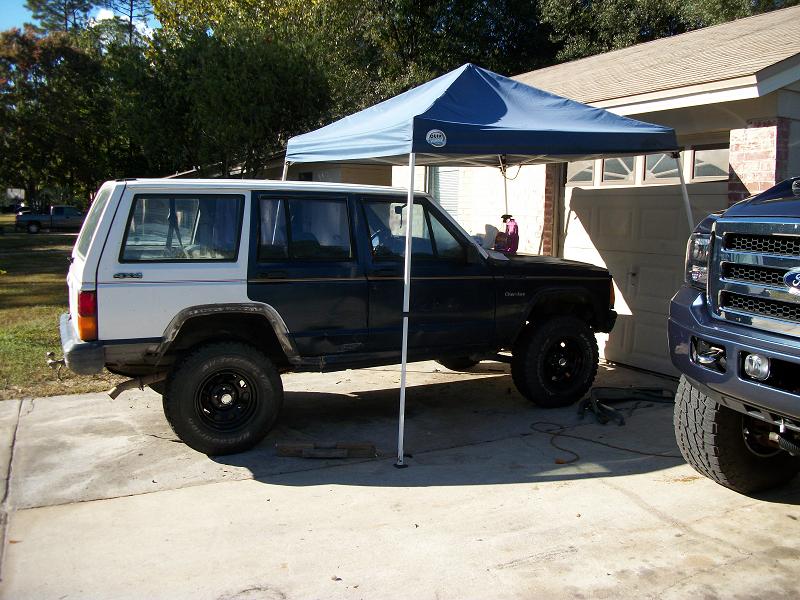 Two-tone Jeep Cherokee with a blue canopy over its side, parked next to a blue Ford truck