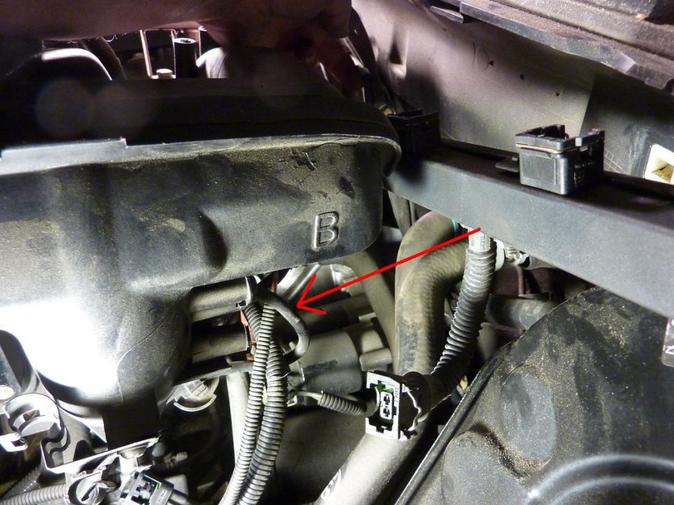 Close-up of engine bay with hoses, wires, and a black plastic cover with a 'B' on it. A red arrow points to a wire harness