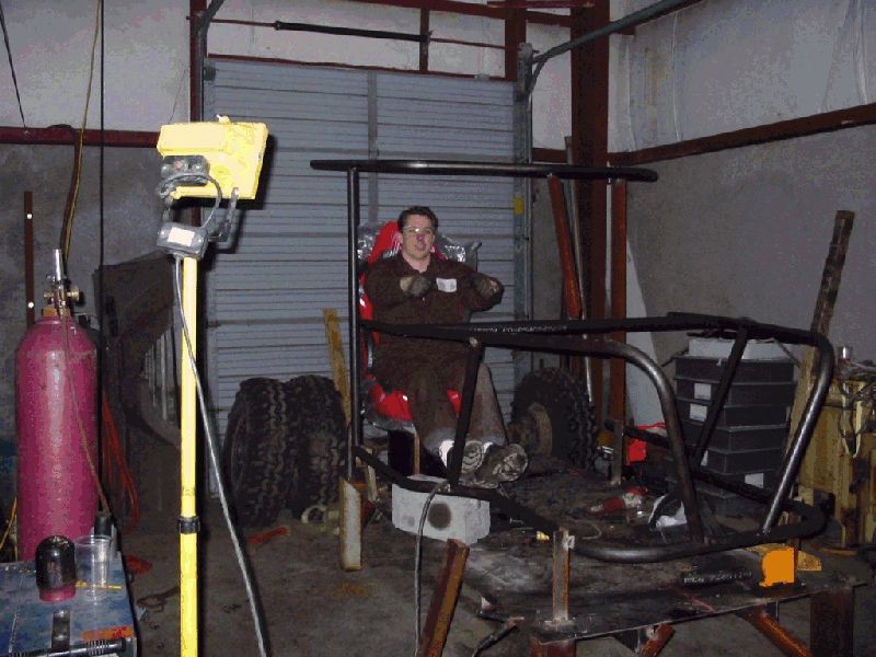 Man seated in red racing seat inside a partially welded tube chassis buggy frame in a shop