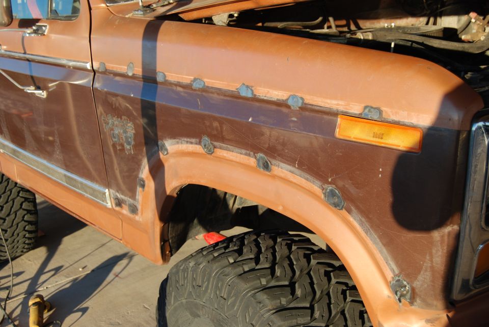Side view of a brown Ford truck fender with rust spots and new welds. Hood is open, showing engine parts