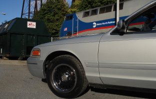 2000 Ford Crown Victoria - A silver sedan is parked next to a dumpster, with a Metro-North Railroad train passing behind it.