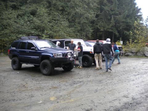 2005 Jeep Wrangler - Several off-road vehicles are gathered in a dirt lot with trees in the background.