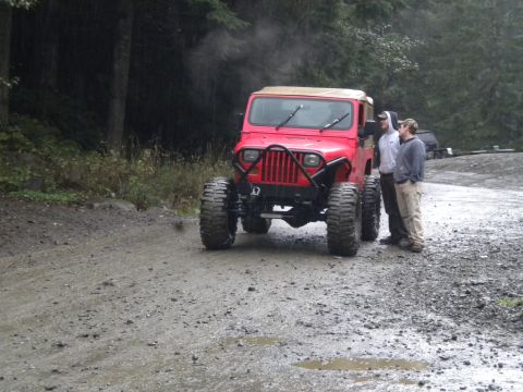2005 Jeep Wrangler - A red Jeep Wrangler sits on a wet, muddy road in a wooded area, with two individuals conversing next to it.