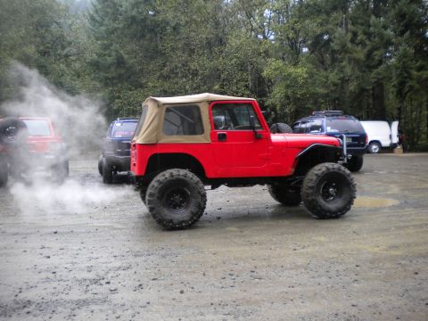 2005 Jeep Wrangler - A red 2005 Jeep Wrangler with a tan soft top is parked on a gravel lot, emitting smoke from its exhaust.