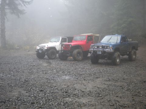 2005 Jeep Wrangler - Three off-road vehicles, including two Jeeps and a pickup truck, are parked in a foggy, wooded area.