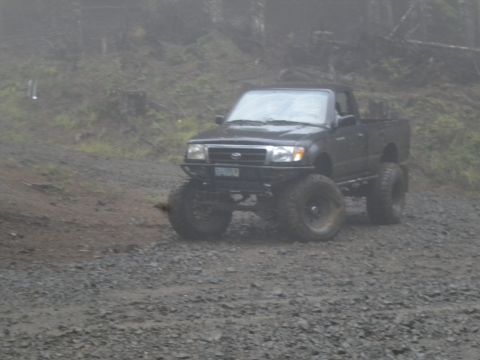 2005 Jeep Wrangler - This lifted pickup truck navigates a rough gravel road in a foggy, wooded environment.