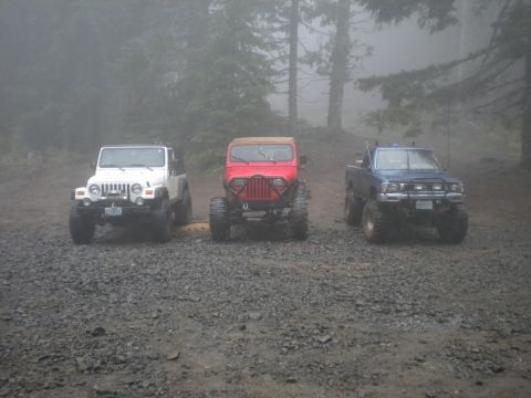 2005 Jeep Wrangler - Three modified off-road vehicles, including a 2005 Jeep Wrangler, parked together on a gravel road.