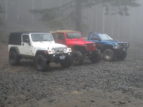 2005 Jeep Wrangler - Three modified off-road vehicles are parked on a gravel surface with trees in the background on a foggy day.