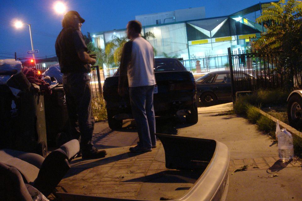 Two men stand by the rear of a dark sedan. Another car is parked behind it