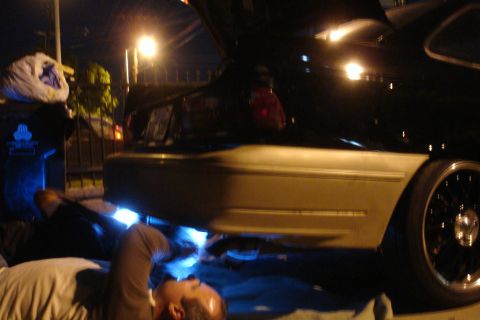 1993 Ford Taurus - A person works on the underside of a black car at night, with the trunk open.